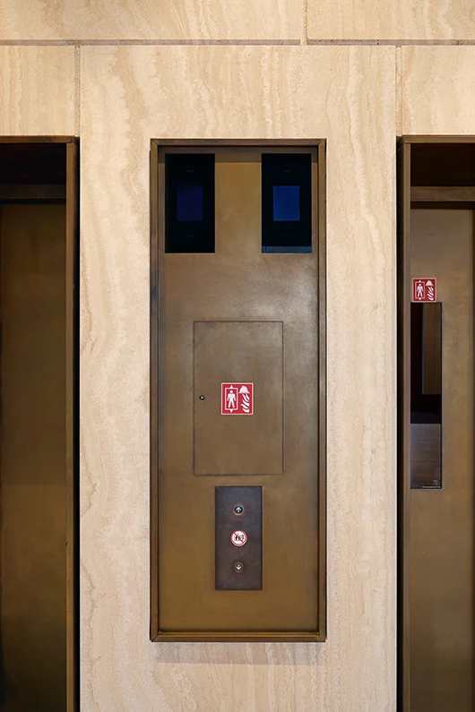Detailaufnahme im Stil einer Architekturfotografie von einem Fahrstuhl und Bedientableau aus Bronze vor braunem Naturstein.